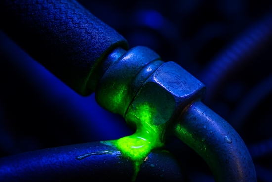 Auto AC Leak Detection Basics ac dye test image - Auto Repair San Antonio | Auto Service Experts Macro photo of automotive AC hose connection showing fluorescent green leak under UV light, with crisp detail of oxidized metal, textured rubber, moisture droplets, and mechanic fingerprint, emphasizing real-world wear and the importance of leak detection.